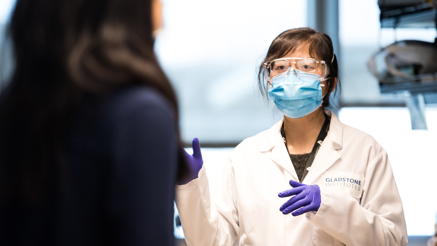 Scientist suited up, wearing a face mask, talking to someone else in the lab