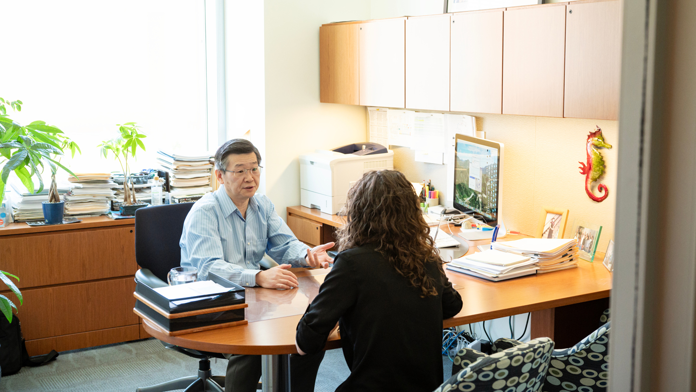 Gladlstone Investigator Yadong Huang sitting at his desk speaking with another scientist