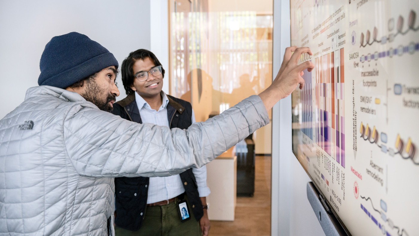 Gladstone Senior Investigator Vijay Ramani (left) and collaborator Siva Kasinathan talking in front of a TV screen with scientific data on it. 